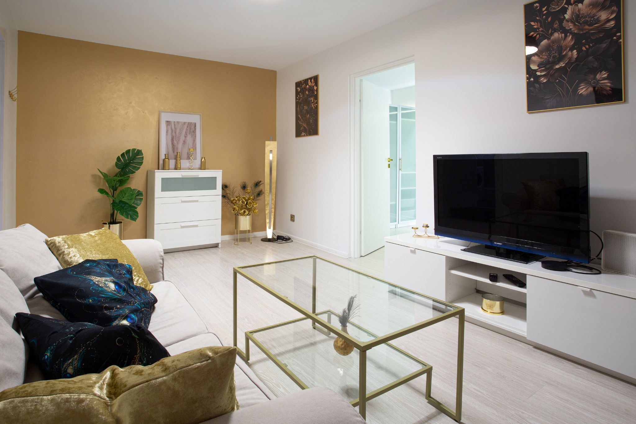 Jasne wnętrze salonu z akcentami złota: sofa z poduszkami, szklany stolik kawowy, złota ściana i dekoracje.