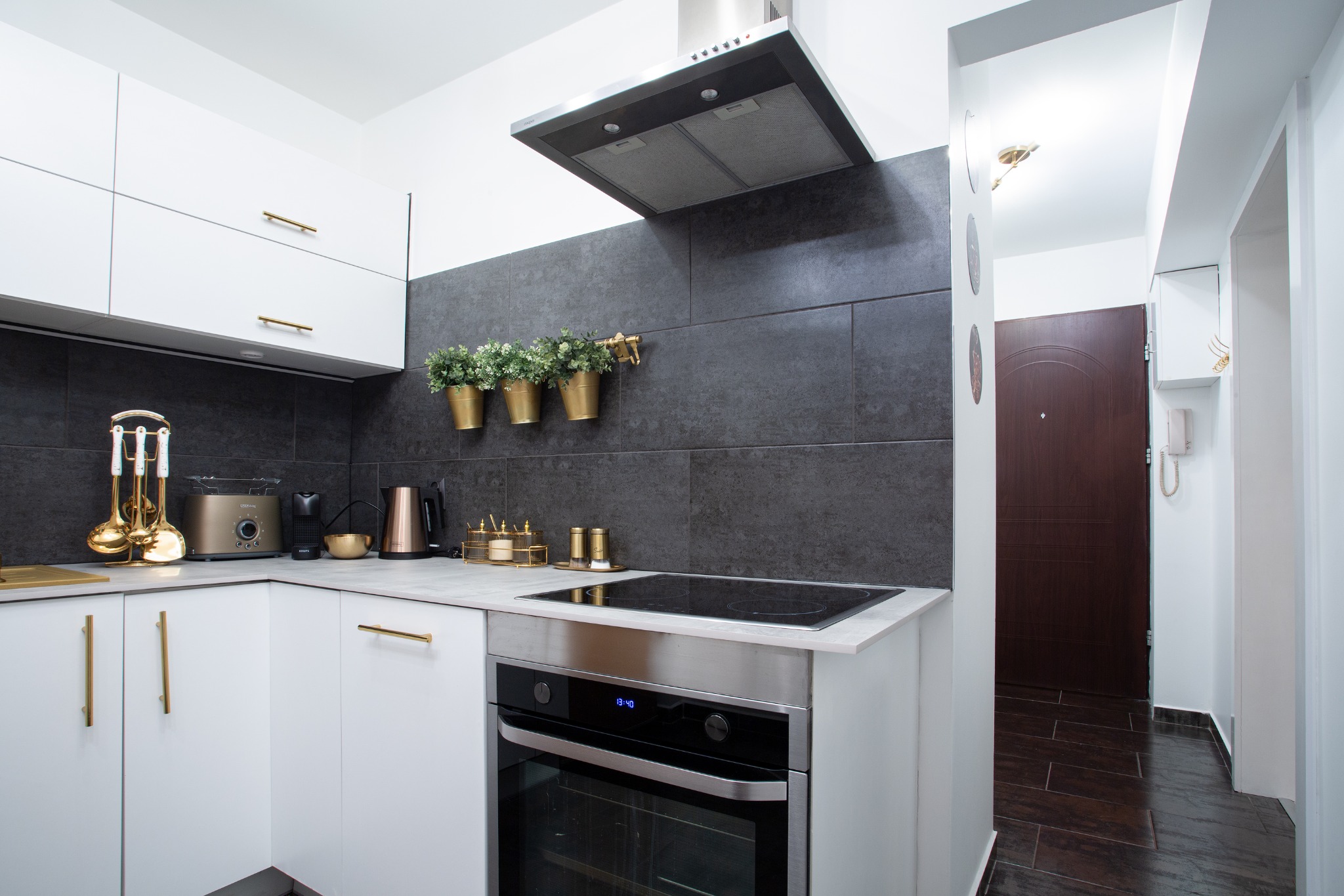 Nowoczesna kuchnia z białymi szafkami, złotymi uchwytami i czarną ścianą z trzema złotymi doniczkami z zielonymi roślinami. Na blacie złoty toster, ekspres do kawy i zestaw przyborów kuchennych.