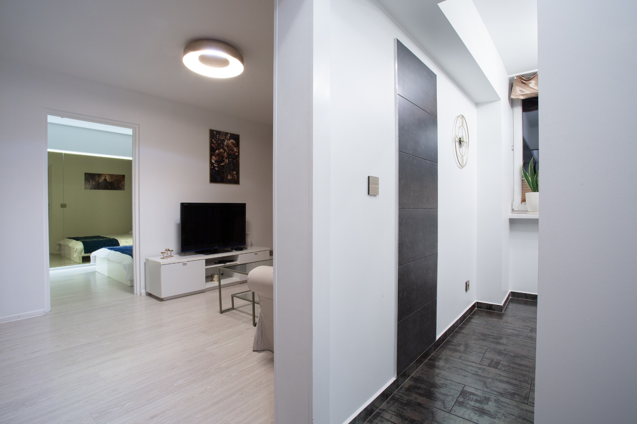 Nowoczesne wnętrze mieszkania z widokiem na sypialnię, jasne panele podłogowe, telewizor na szafce RTV, dekoracyjna ściana z ciemnymi płytami, okrągły zegar na ścianie.