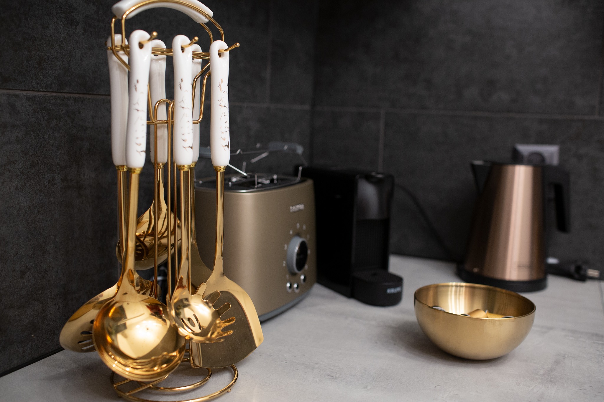 Zestaw złotych przyborów kuchennych na stojaku z porcelanowymi uchwytami, w tle toster, ekspres do kawy i czajnik w odcieniach złota i czerni na blacie z szarego kamienia.