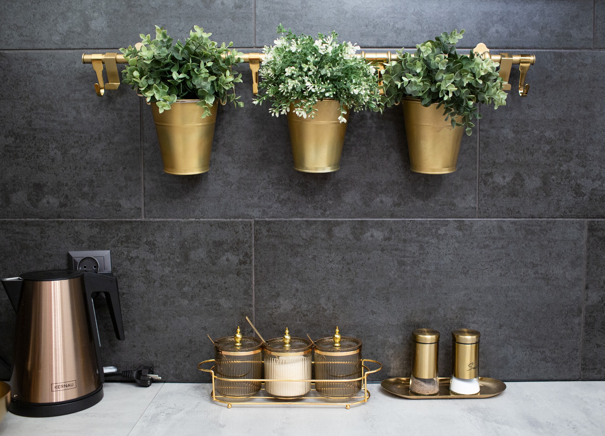Kuchnia z szarymi płytkami, złotymi doniczkami z roślinami i złotymi akcesoriami kuchennymi, czajnik elektryczny Kernau po lewej stronie.