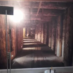Wnętrze pokoju z fototapetą przedstawiającą kopalniany tunel z drewnianą konstrukcją, widoczna lampa stojąca i fragment ściany z gniazdkiem elektrycznym.