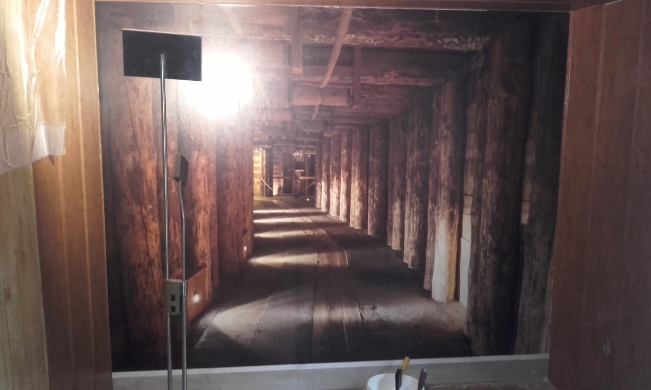 Wnętrze pokoju z fototapetą przedstawiającą kopalniany tunel z drewnianą konstrukcją, widoczna lampa stojąca i fragment ściany z gniazdkiem elektrycznym.