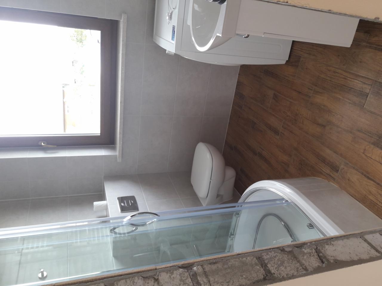 Ukończona łazienka z drewnopodobnymi panelami na podłodze, szarą glazurą ścienną, kabiną prysznicową z przesuwnymi drzwiami, podwieszaną toaletą i oknem z brązową ramą.