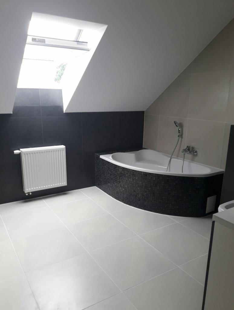 Wykończona łazienka na poddaszu z czarno-białą kolorystyką, widoczna wanna obudowana czarną mozaiką, biały grzejnik na czarnej ścianie oraz okno dachowe.