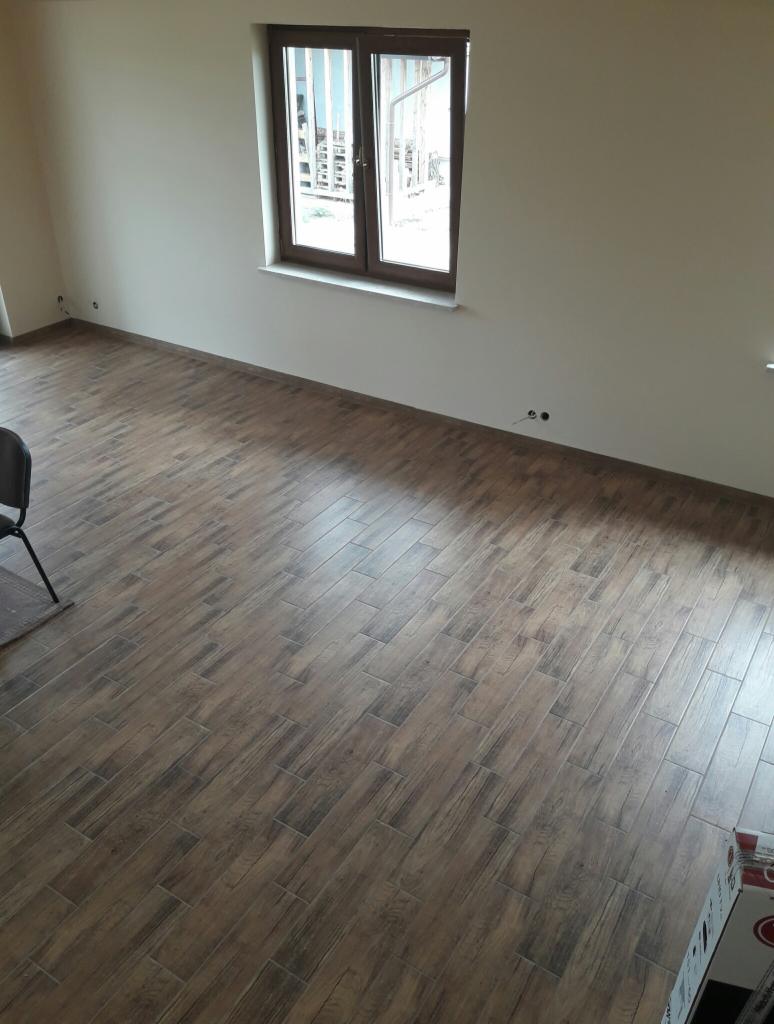 Ukończone pomieszczenie z brązowymi panelami podłogowymi imitującymi drewno, jasne ściany, okno w brązowej ramie i wystające kable elektryczne ze ściany.