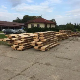 Stosy świeżo przetartej tarcicy z korą ułożone na betonowym placu, w tle budynki gospodarcze i zaparkowane samochody osobowe.