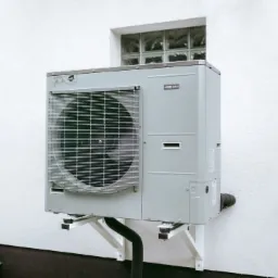 Zewnętrzna jednostka pompy ciepła zamontowana na białej ścianie budynku, widoczna kratka wentylacyjna i rury odprowadzające, na tle okno z luksferami.