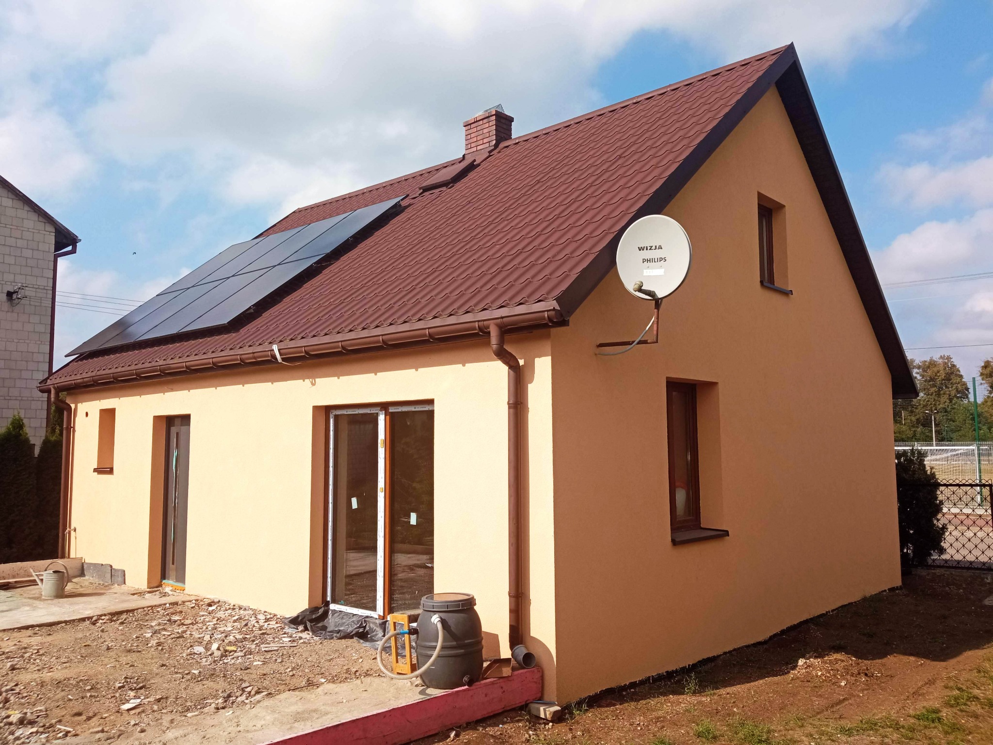 Niski dom z brązowym dachem i panelami słonecznymi, widoczna antena satelitarna, elewacja w kolorze piaskowym, teren wokół domu w trakcie prac wykończeniowych.