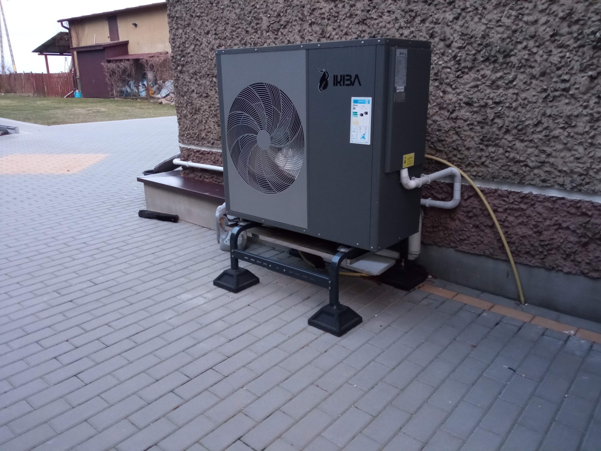 Szara pompa ciepła marki IKIBA zamontowana na metalowym stelażu przy szorstkiej ścianie budynku, widoczne rury doprowadzające.