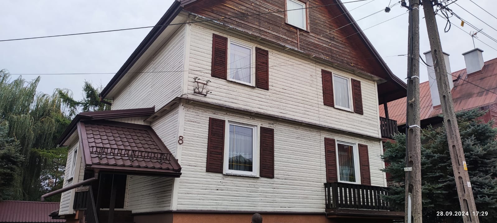 Dwupiętrowy dom z drewnianą elewacją, brązowymi okiennicami i numerem 8. Widoczne słupy energetyczne i fragmenty sąsiednich budynków.