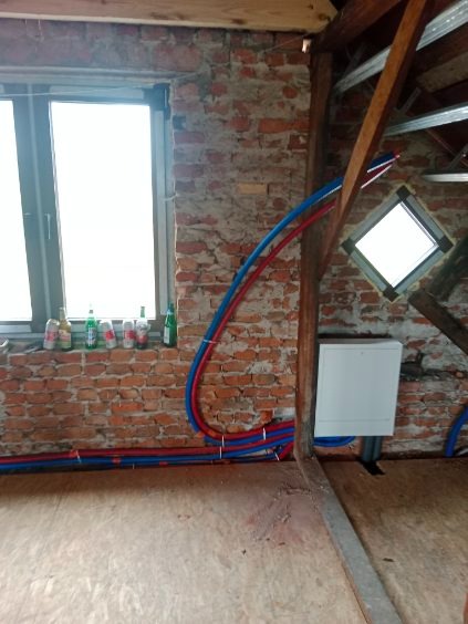 Instalacja CO w surowym wnętrzu poddasza: Czerwono-niebieskie rury grzewcze prowadzone wzdłuż ceglanej ściany i drewnianej konstrukcji dachu. Widoczne okno i biała skrzynka.