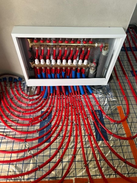 Rozdzielacz ogrzewania podłogowego z czerwonymi rurkami na srebrnej macie izolacyjnej. Widoczne liczne zawory i złączki. Ujęcie z góry, blisko ściany.