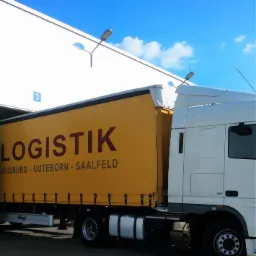 Żółta naczepa ciężarówki z napisem 'LOGISTIK' i nazwami miast Duisburg, Guteborn, Saalfeld, zaparkowana przy rampie magazynowej w słoneczny dzień.