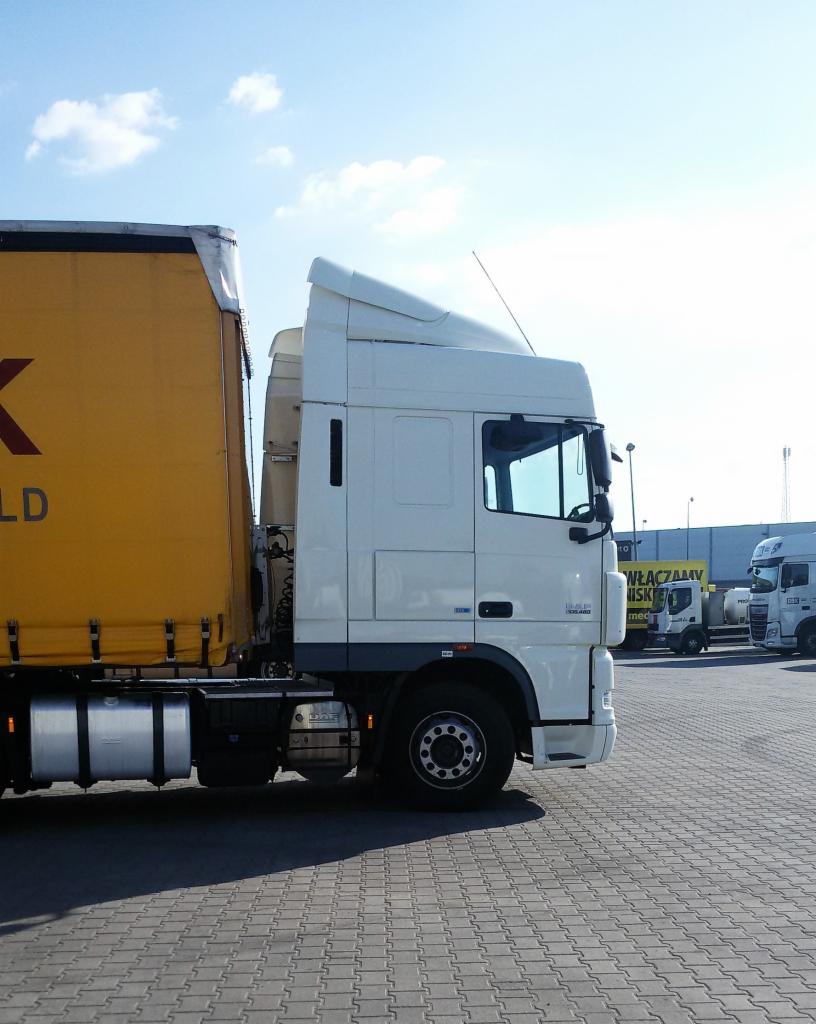 Ciężarówka z żółtą naczepą na brukowanym parkingu, widok z boku kabiny w słoneczny dzień.