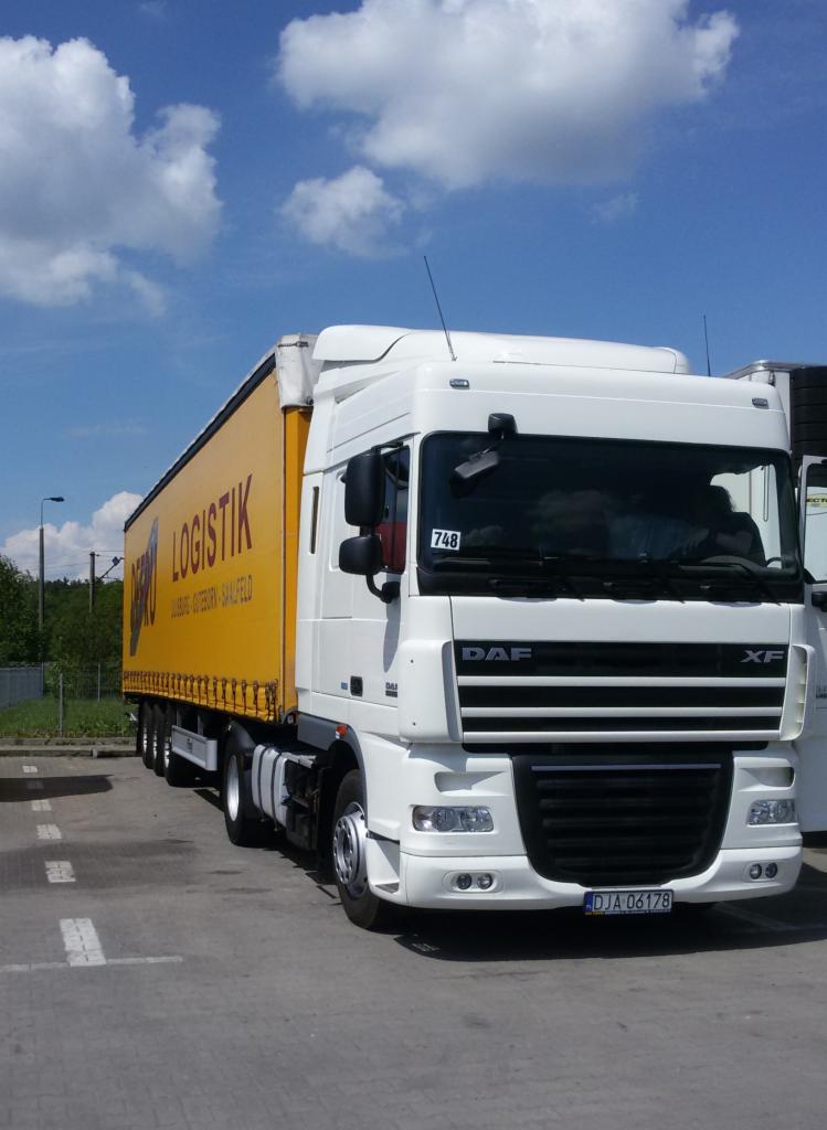 Biała ciężarówka DAF XF z żółtą naczepą LOGISTIK, zaparkowana na parkingu w słoneczny dzień.