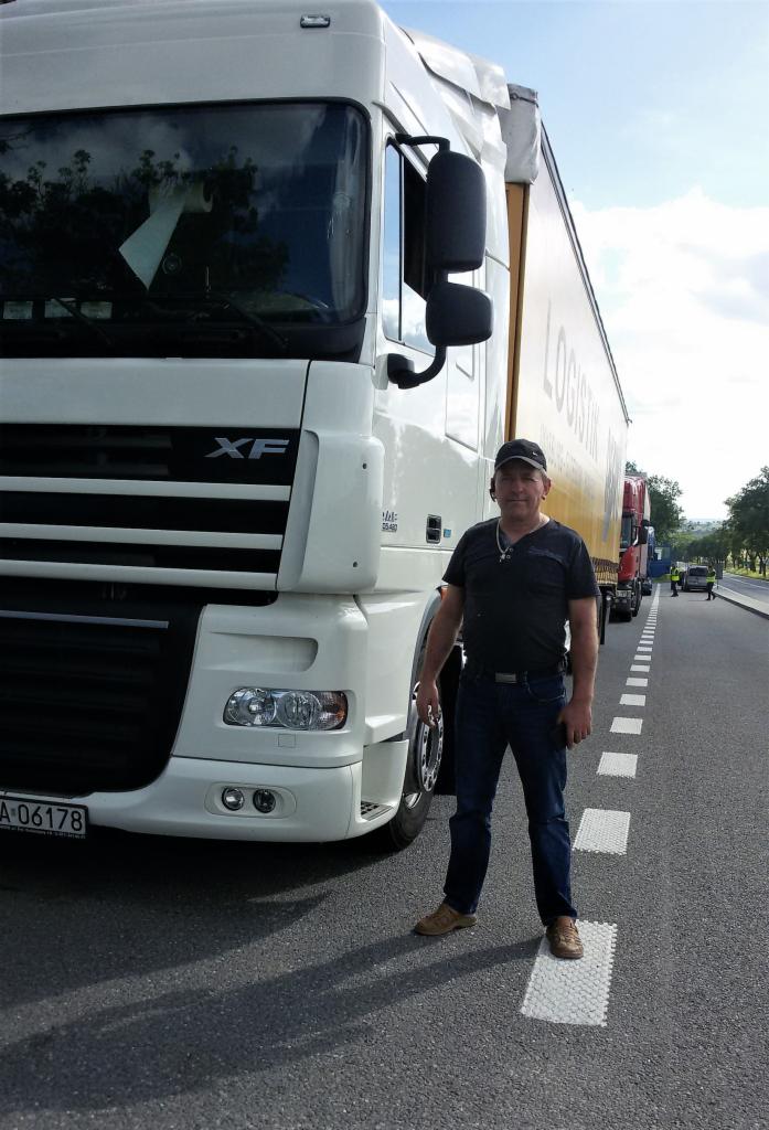 Kierowca w czarnej koszulce i dżinsach stoi przed białym ciągnikiem siodłowym DAF XF z naczepą, na tle drogi z przerywaną linią.