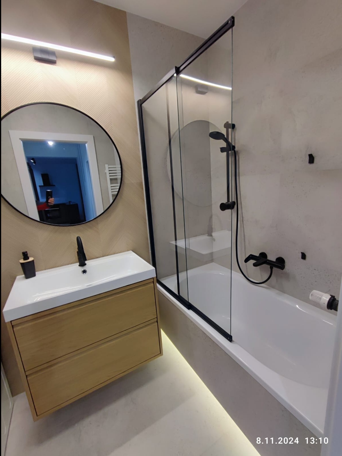 Nowoczesna łazienka z kabiną prysznicową w czarnej ramie, umywalką z drewnianą szafką i okrągłym lustrem, oświetlona listwą LED pod wanną.