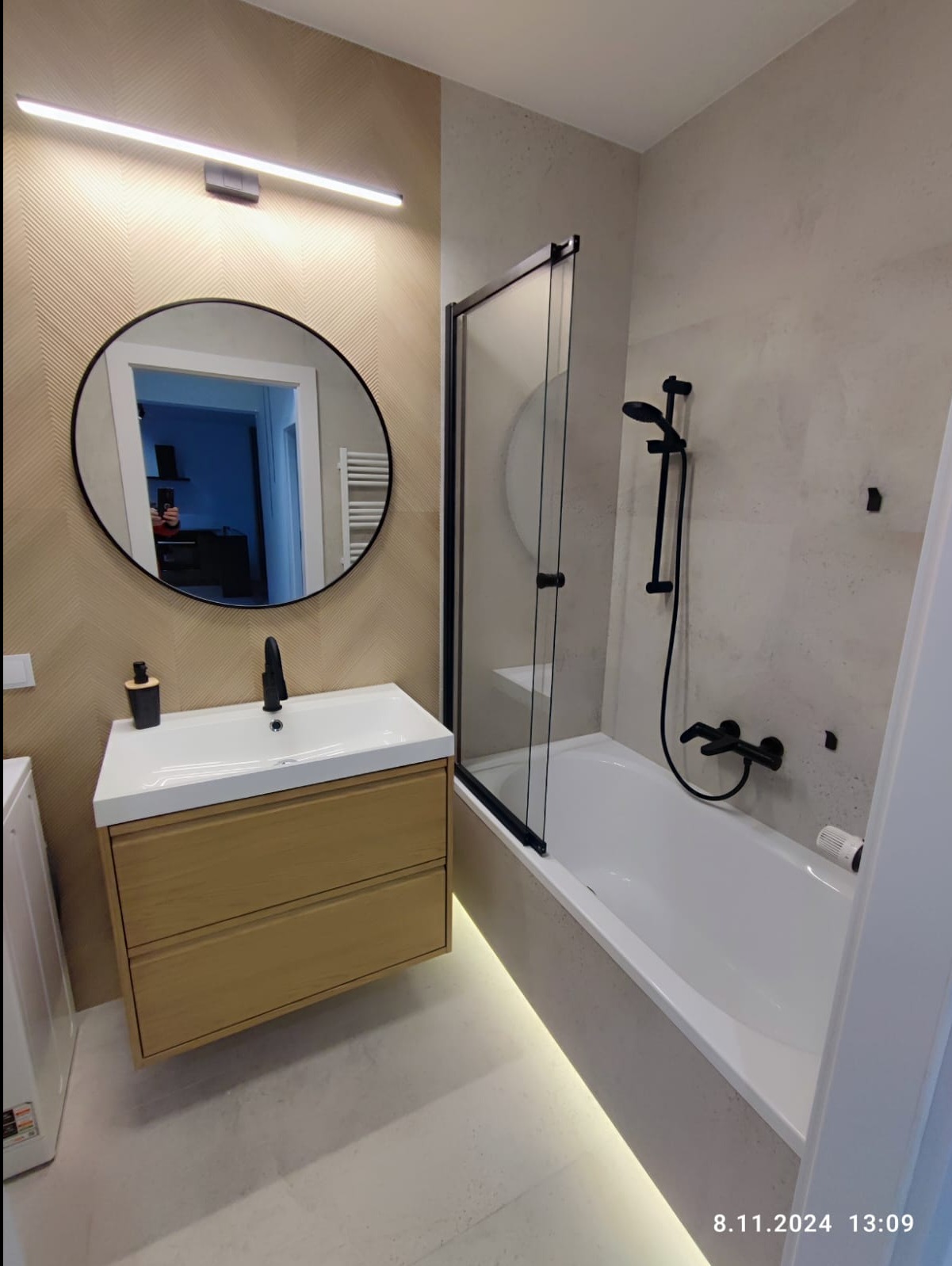 Nowoczesna łazienka z okrągłym lustrem, drewnianą szafką pod umywalką, czarną armaturą i wanną z parawanem, podświetlona listwą LED przy podłodze.