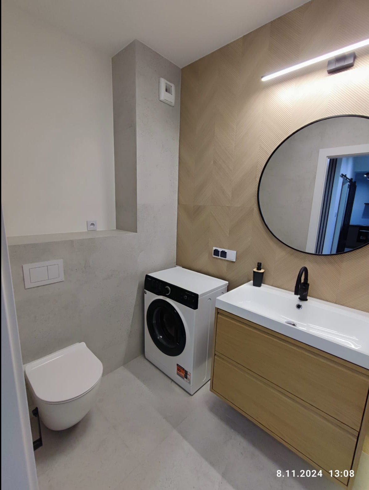 Nowoczesna łazienka z toaletą, pralką i umywalką z szafką w kolorze drewna, okrągłe lustro z czarną ramą, ściana wykończona płytkami w geometryczny wzór jodełki.