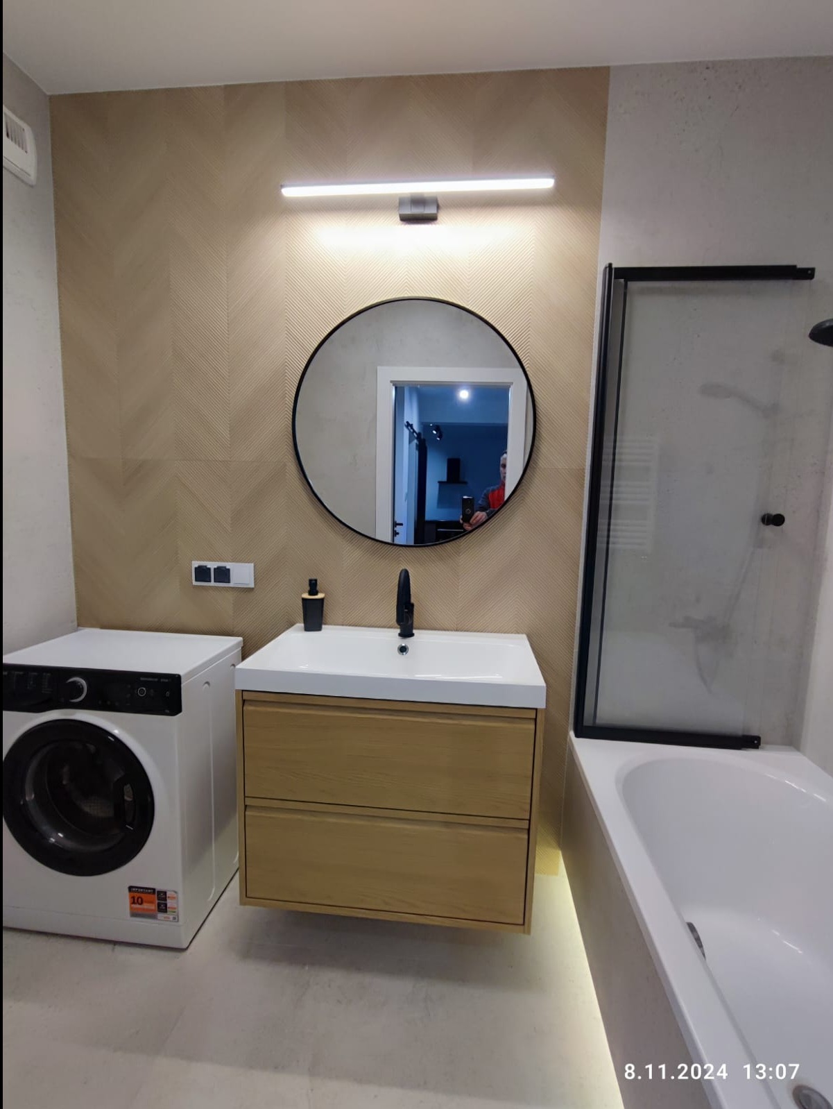 Nowoczesna łazienka z pralką, umywalką z szafką z jasnego drewna, okrągłym lustrem i kabiną prysznicową z czarnymi ramami; beżowe płytki na ścianie ułożone w jodełkę.