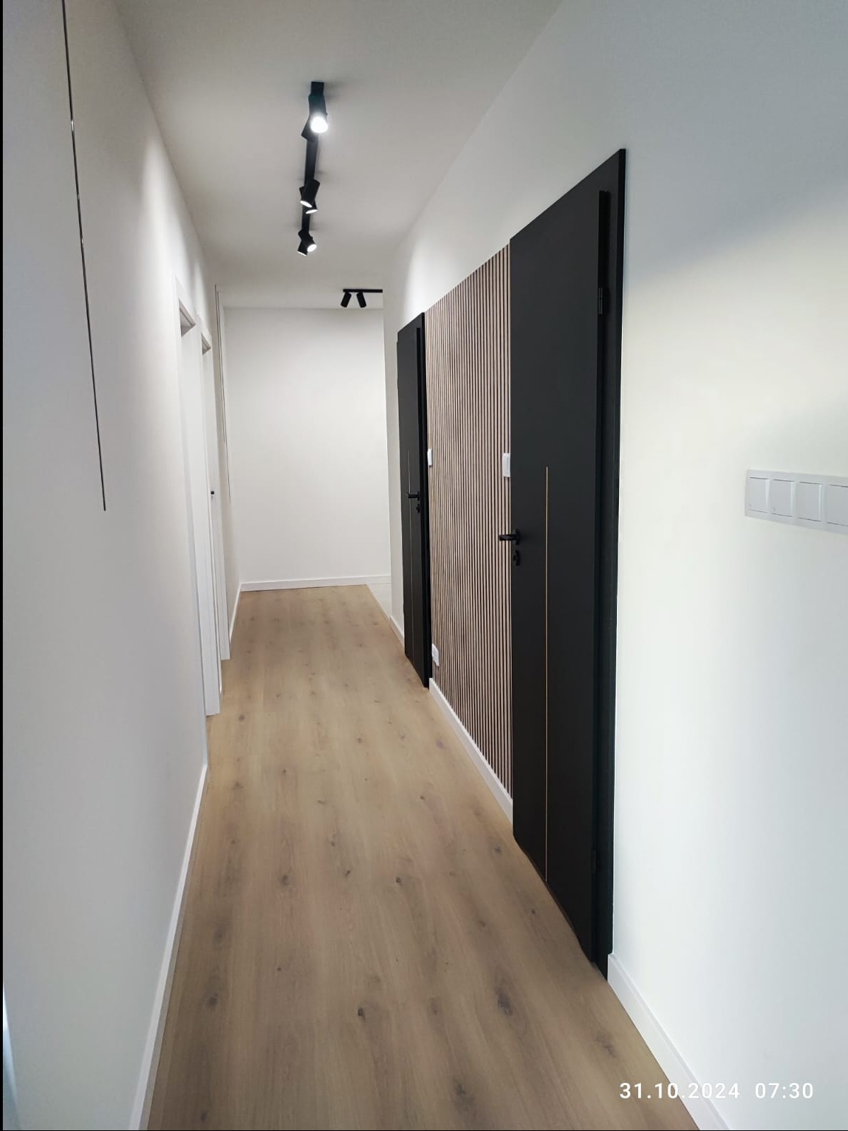 Nowoczesny korytarz z drewnianą podłogą, białymi ścianami i czarnymi drzwiami z drewnianymi lamelami, oświetlony reflektorami na szynie.