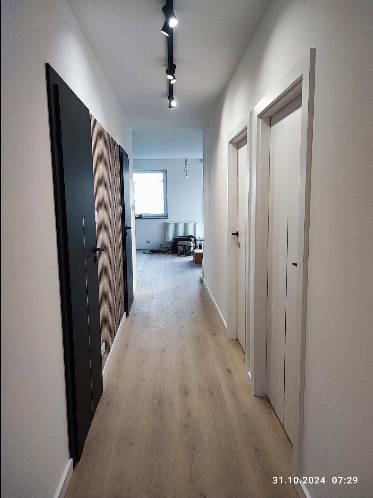 Nowoczesny korytarz po remoncie z drewnianą podłogą, panelami lamelowymi na ścianie i minimalistycznymi drzwiami w kolorze białym i czarnym, oświetlony reflektorami sufitowymi.