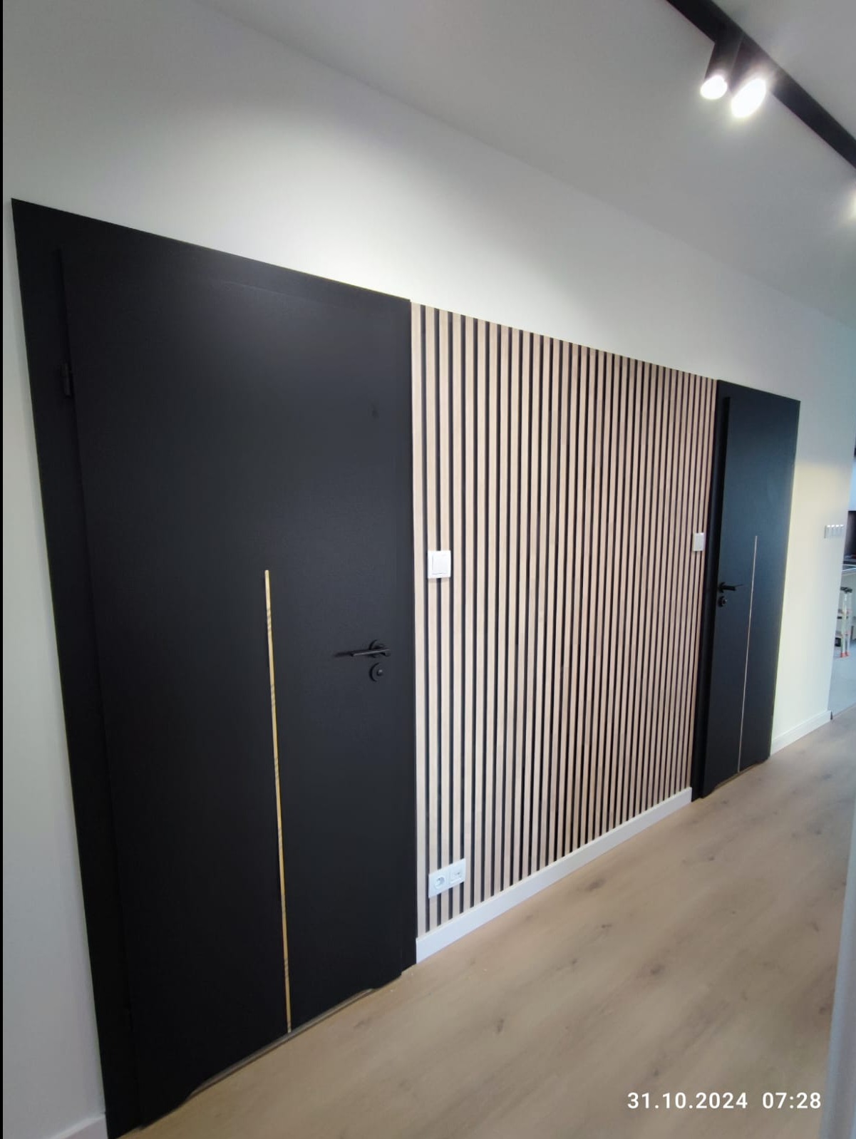 Nowoczesna aranżacja ściany w Gorzowie Wielkopolskim z czarnymi drzwiami i dekoracyjnymi panelami pionowymi z jasnego drewna, tworzącymi rytmiczny wzór.