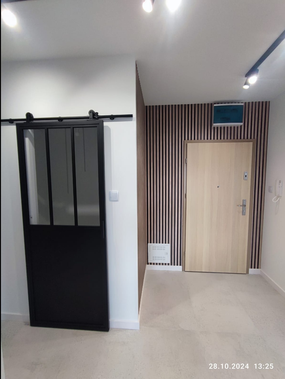 Nowoczesne wnętrze przedpokoju z przesuwnymi drzwiami loftowymi na szynie, drewnianą okładziną ścienną z pionowych lameli i jasną podłogą z płytek.