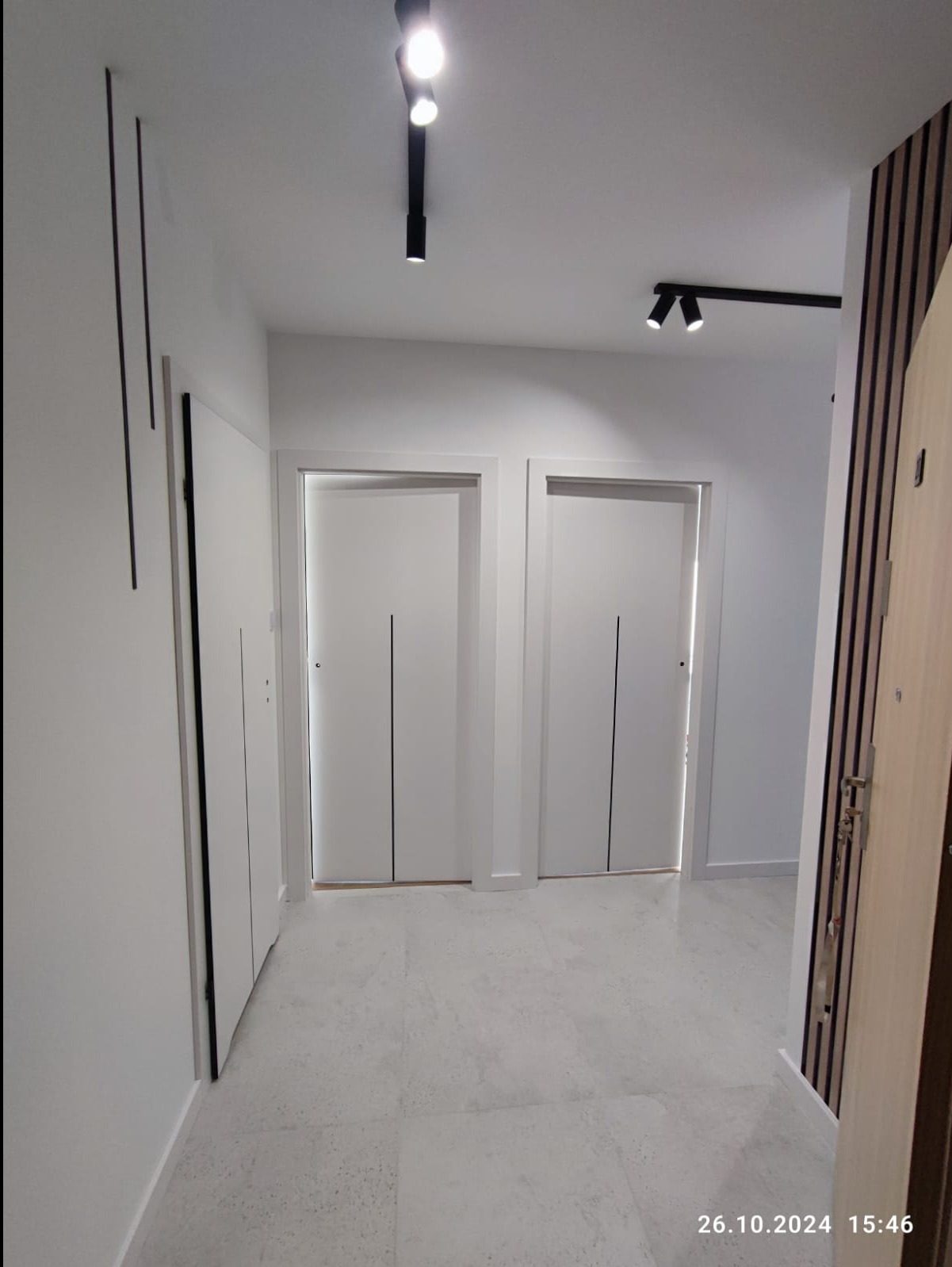 Wykończony korytarz w jasnych barwach z dwiema parami białych drzwi z czarnymi pionowymi akcentami, oświetleniem punktowym na suficie i marmurową podłogą.