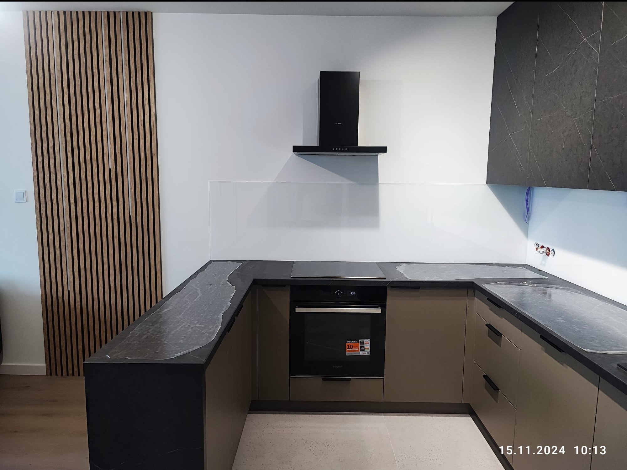 Nowoczesna kuchnia z brązowymi szafkami, blatem z ciemnego marmuru z białymi żyłami, drewnianą ścianką z pionowych listew i czarnym okapem, zabezpieczona folią ochronną na blacie.