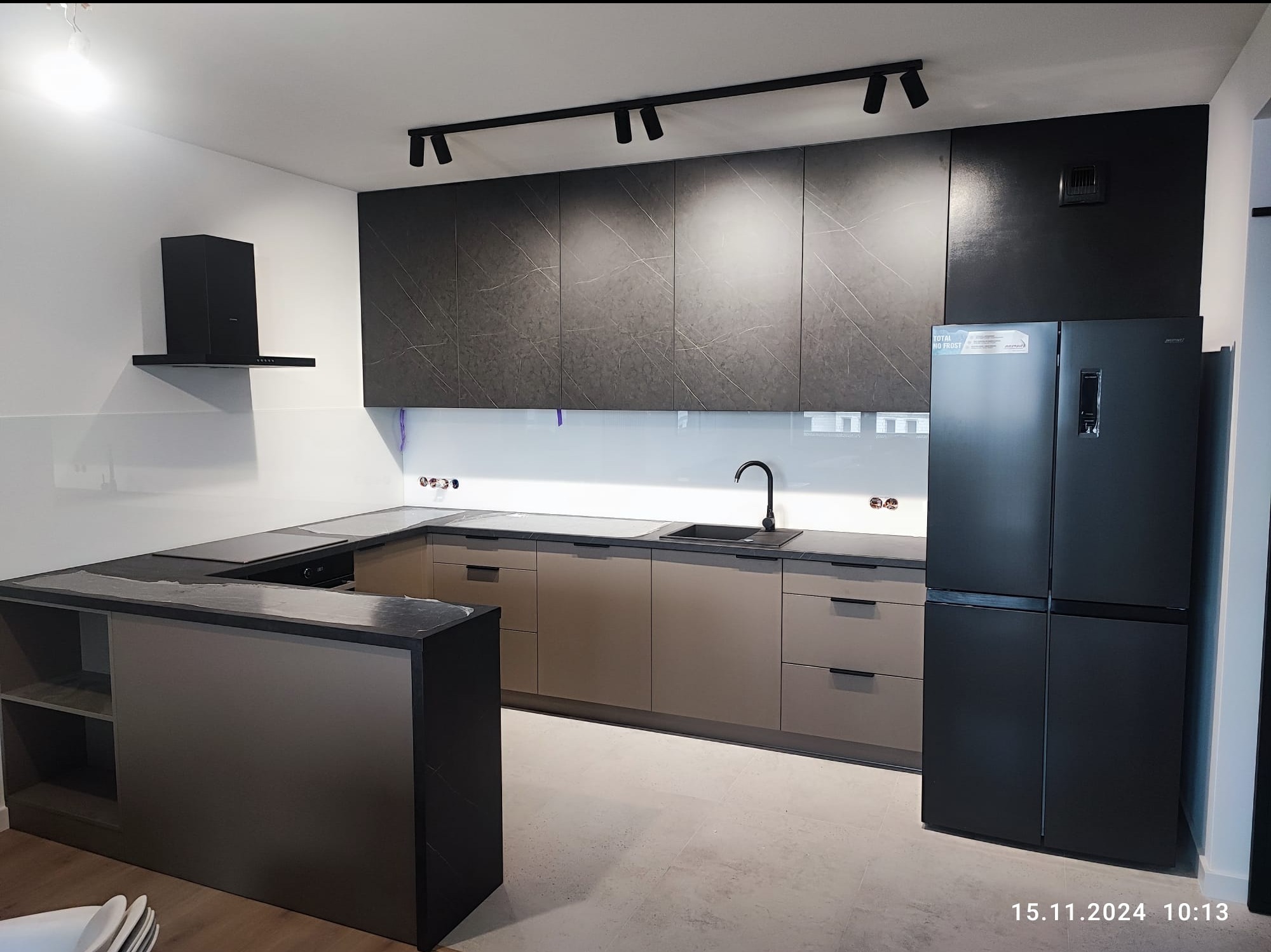 Nowoczesna kuchnia z szarymi szafkami z marmurowym wzorem, czarnym blatem i wyspą, oświetleniem punktowym na suficie i czarną lodówką z datą 15.11.2024 w rogu.