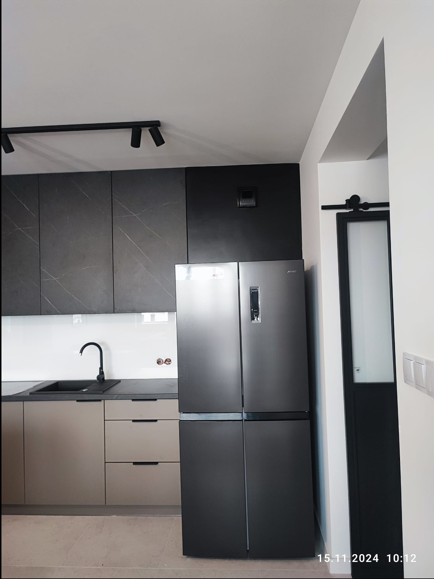 Nowoczesna kuchnia z czarnymi szafkami górnymi z marmurowym wzorem, beżowymi szafkami dolnymi i czarną armaturą, oświetlona reflektorami, z widoczną czarną przesuwną szklaną ścianą w tle.