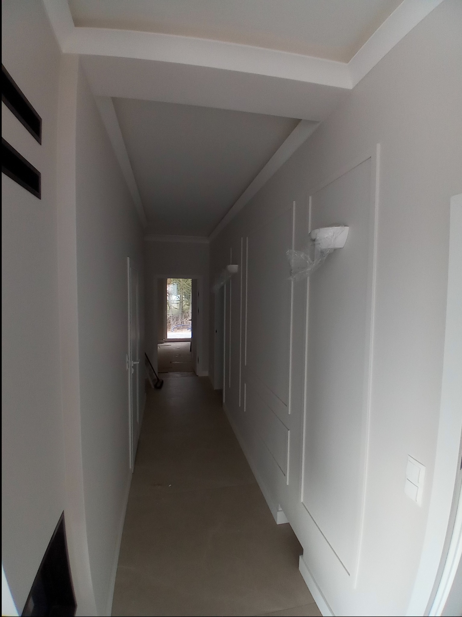 Biały korytarz z panelami ściennymi i listwami przysufitowymi, widoczne lampy ścienne zabezpieczone folią i surowe wykończenie podłogi.