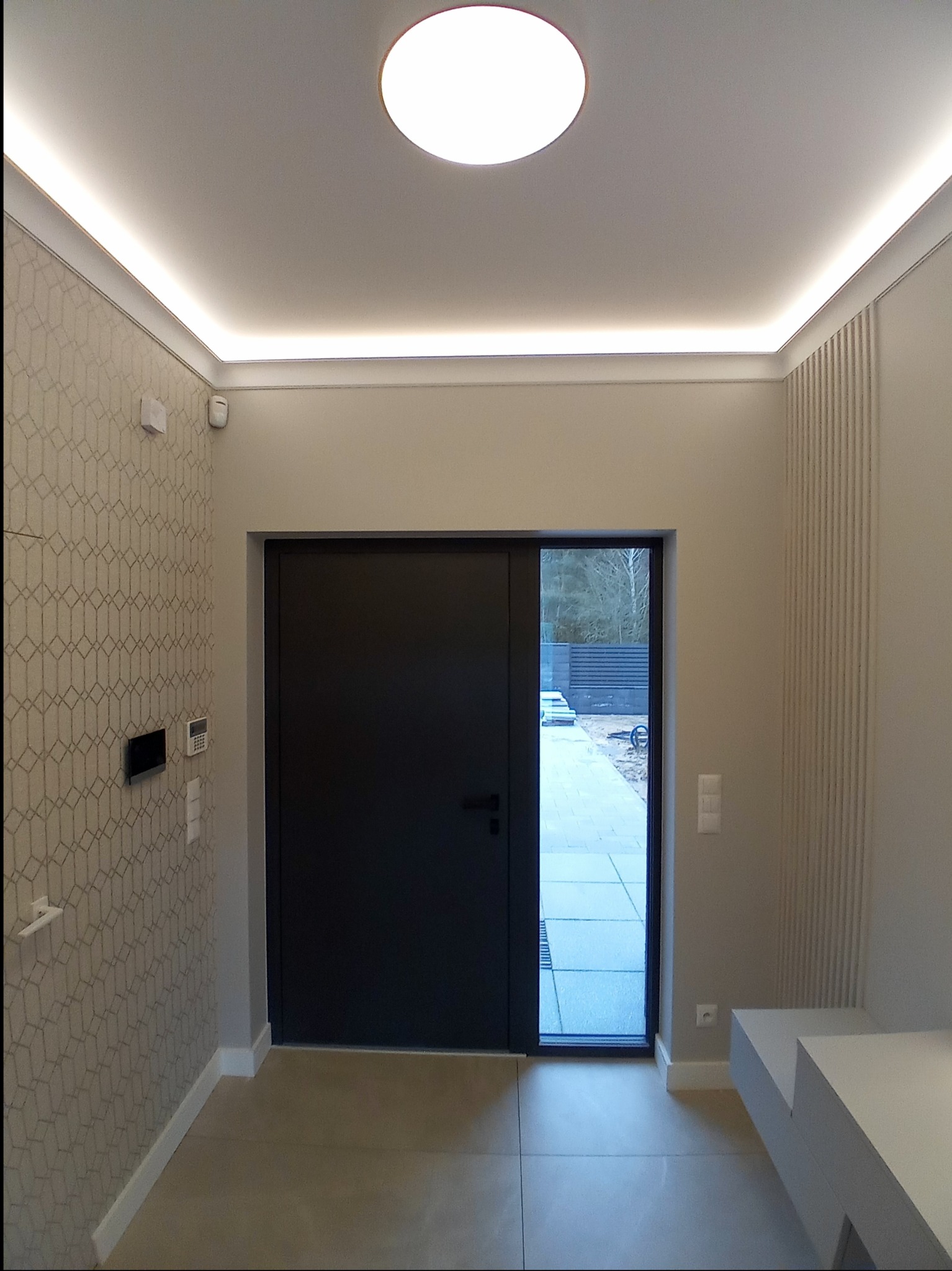 Nowoczesny przedpokój z geometryczną tapetą, ukrytym oświetleniem LED w suficie i nad drzwiami, oraz minimalistycznym siedziskiem, widok na zewnątrz przez przeszkloną część drzwi.