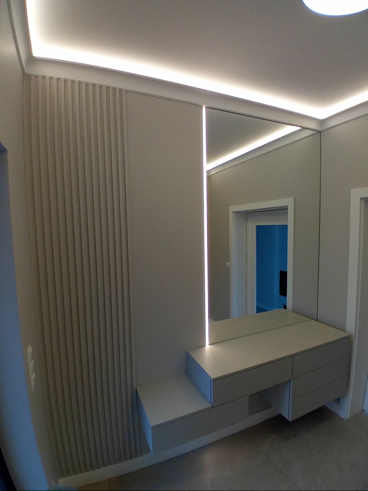 Nowoczesna aranżacja przedpokoju z lustrem z podświetleniem LED, pionowymi panelami dekoracyjnymi na ścianie i minimalistyczną zabudową meblową w odcieniach szarości.