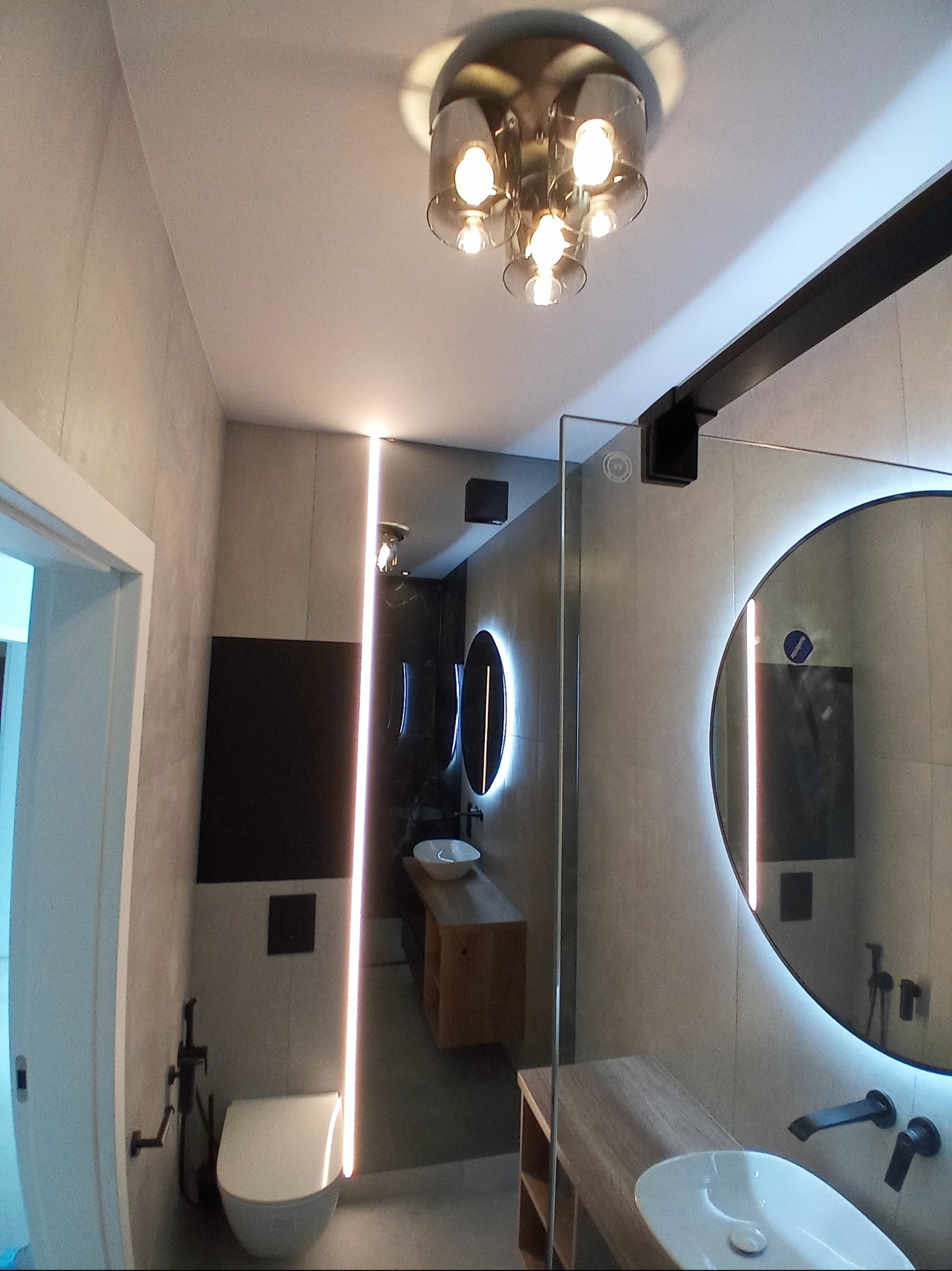 Nowoczesna łazienka z podświetlanymi lustrami, szklaną kabiną prysznicową i designerską lampą sufitową, widok z perspektywy wejścia.