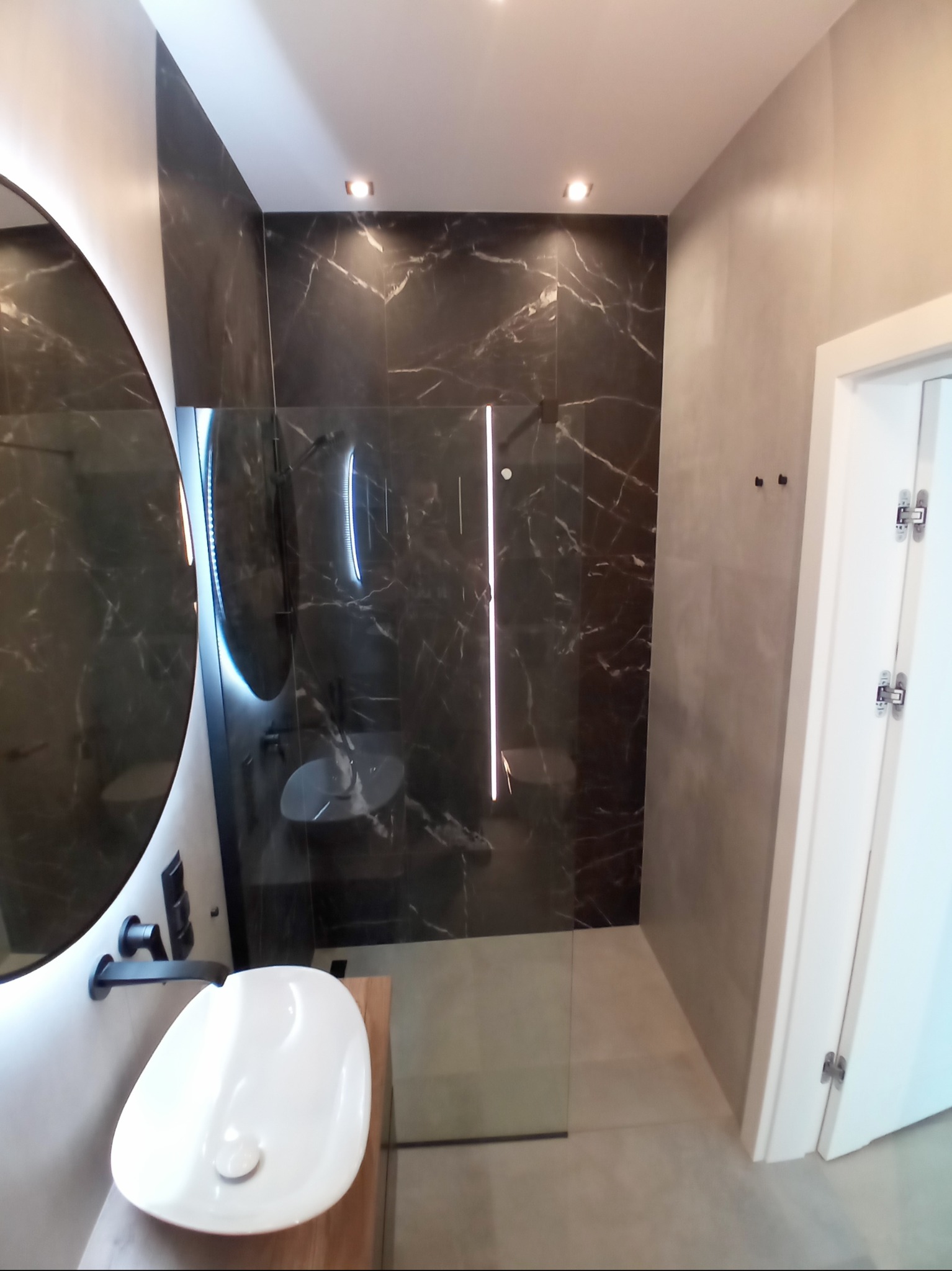 Nowoczesna łazienka z czarnymi marmurowymi płytkami, okrągłym lustrem z podświetleniem LED, białą umywalką nablatową i szklaną kabiną prysznicową.