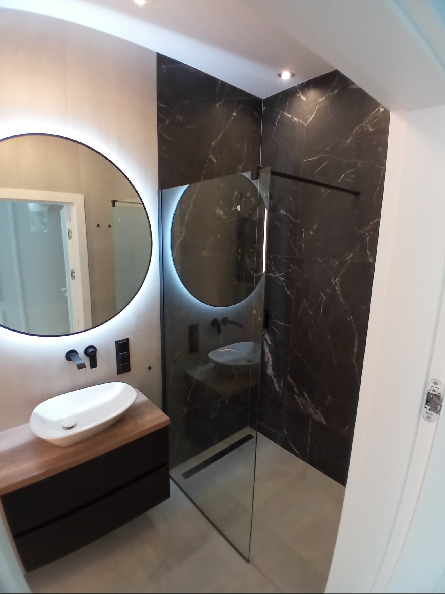 Nowoczesna łazienka z okrągłym lustrem LED, czarnymi płytkami imitującymi marmur w strefie prysznica walk-in i drewnianą szafką pod umywalką.