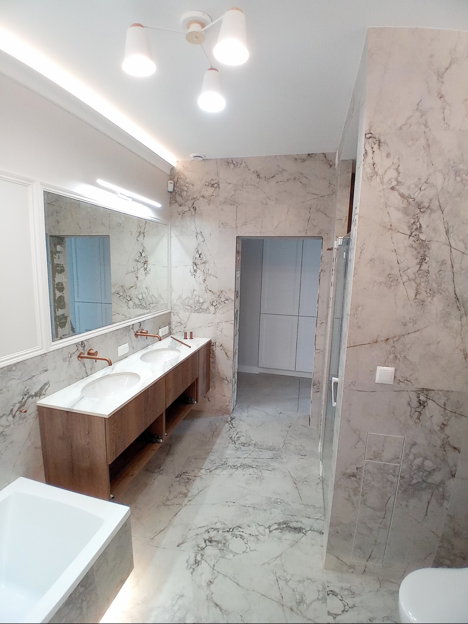 Nowoczesna łazienka z marmurowymi płytkami, podwójną umywalką nablatową z miedzianą armaturą, oświetleniem LED, oraz widoczną kabiną prysznicową i częścią wanny.