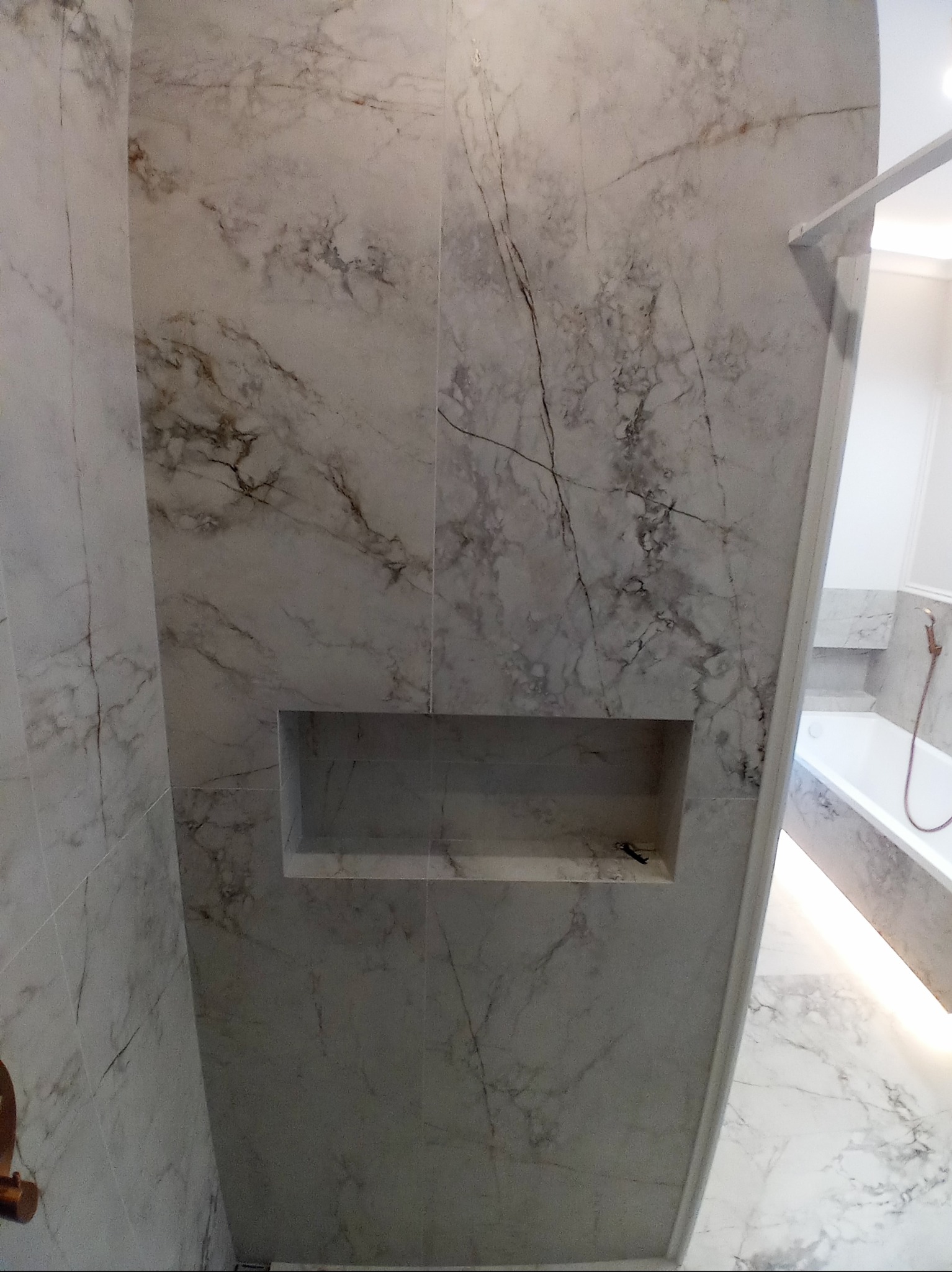 Wykończona łazienka z płytkami imitującymi marmur, wnęka w ścianie wyłożona tym samym materiałem, widoczna wanna i oświetlenie LED.