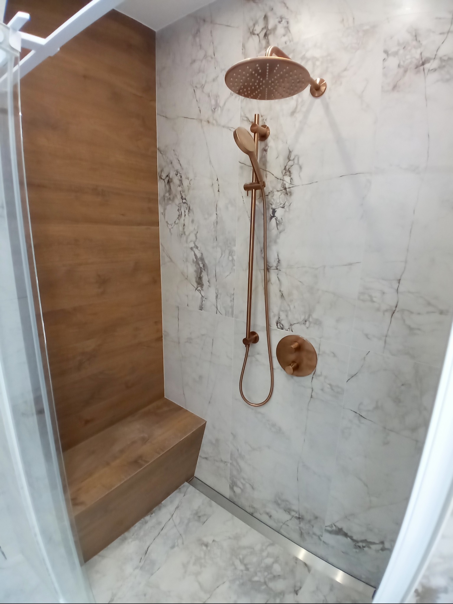 Nowoczesna kabina prysznicowa z siedziskiem wykończona marmurowymi płytkami i armaturą w kolorze różowego złota.