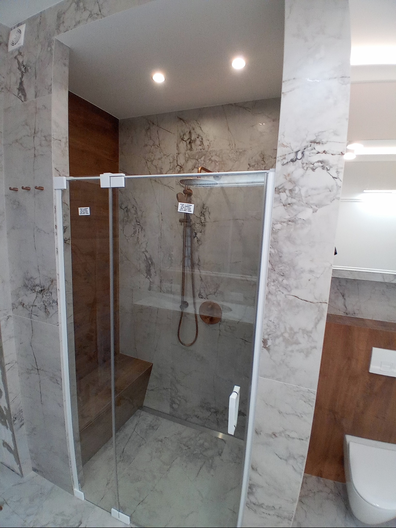 Kabina prysznicowa z siedziskiem, wykończona płytkami imitującymi marmur, z widoczną armaturą w kolorze miedzi i detalami w postaci etykiet informacyjnych na szkle.