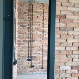 Odsłonięta instalacja elektryczna w peszlach prowadzona po ceglanej ścianie w stylu loftowym, widoczne gniazda elektryczne i czarna rama konstrukcyjna.