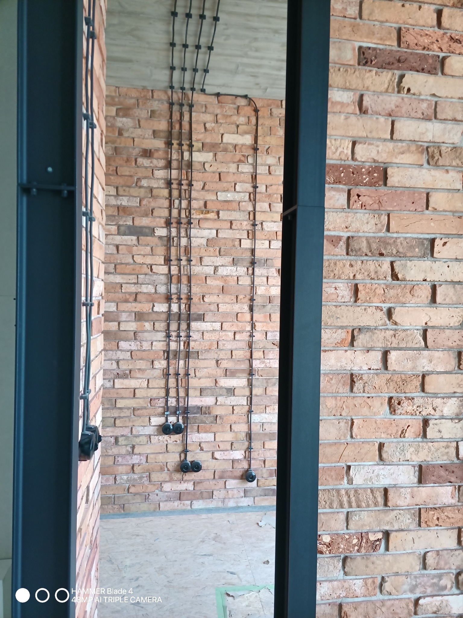 Odsłonięta instalacja elektryczna w peszlach prowadzona po ceglanej ścianie w stylu loftowym, widoczne gniazda elektryczne i czarna rama konstrukcyjna.