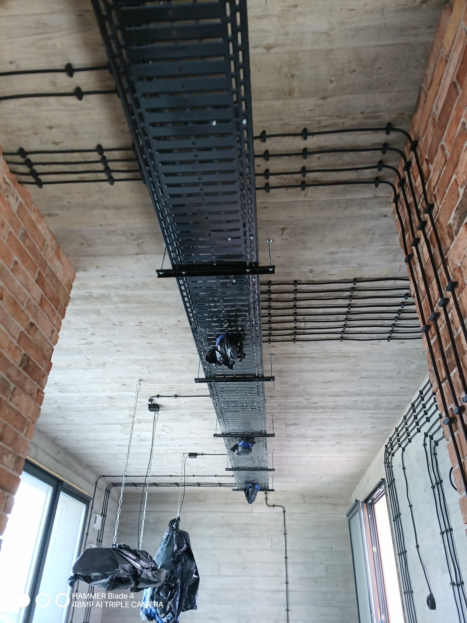Widok z dołu na sufit z betonową fakturą, na którym zamontowano koryto kablowe z czarnymi przewodami elektrycznymi prowadzonymi w peszlach i przymocowanymi do ściany z cegły klinkierowej.