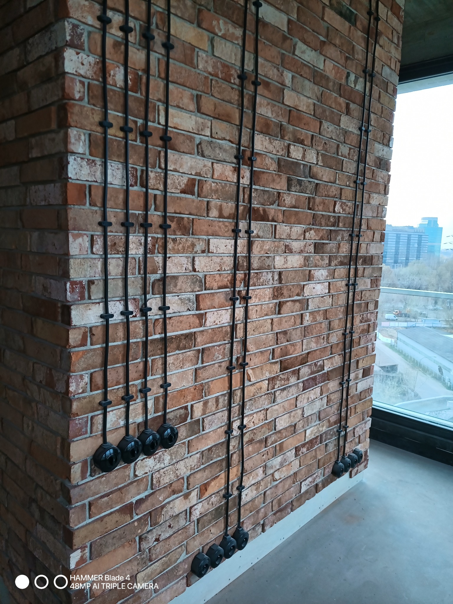 Instalacja elektryczna natynkowa w stylu retro na ceglanej ścianie, widoczne czarne przewody i ceramiczne uchwyty mocujące, z okna widok na miasto.