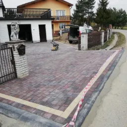 Nowo ułożona, kolorowa kostka brukowa na podjeździe z dekoracyjnym pasem, widoczna zagęszczarka i rzeźba lwa na postumencie przy bramie wjazdowej.
