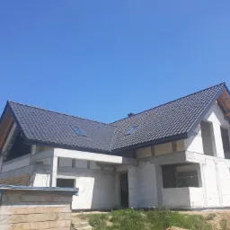 Dom w stanie surowym zamkniętym z grafitową dachówką ceramiczną, widoczne okna dachowe i drewniane elementy więźby dachowej, na pierwszym planie skrzynia z deskami.