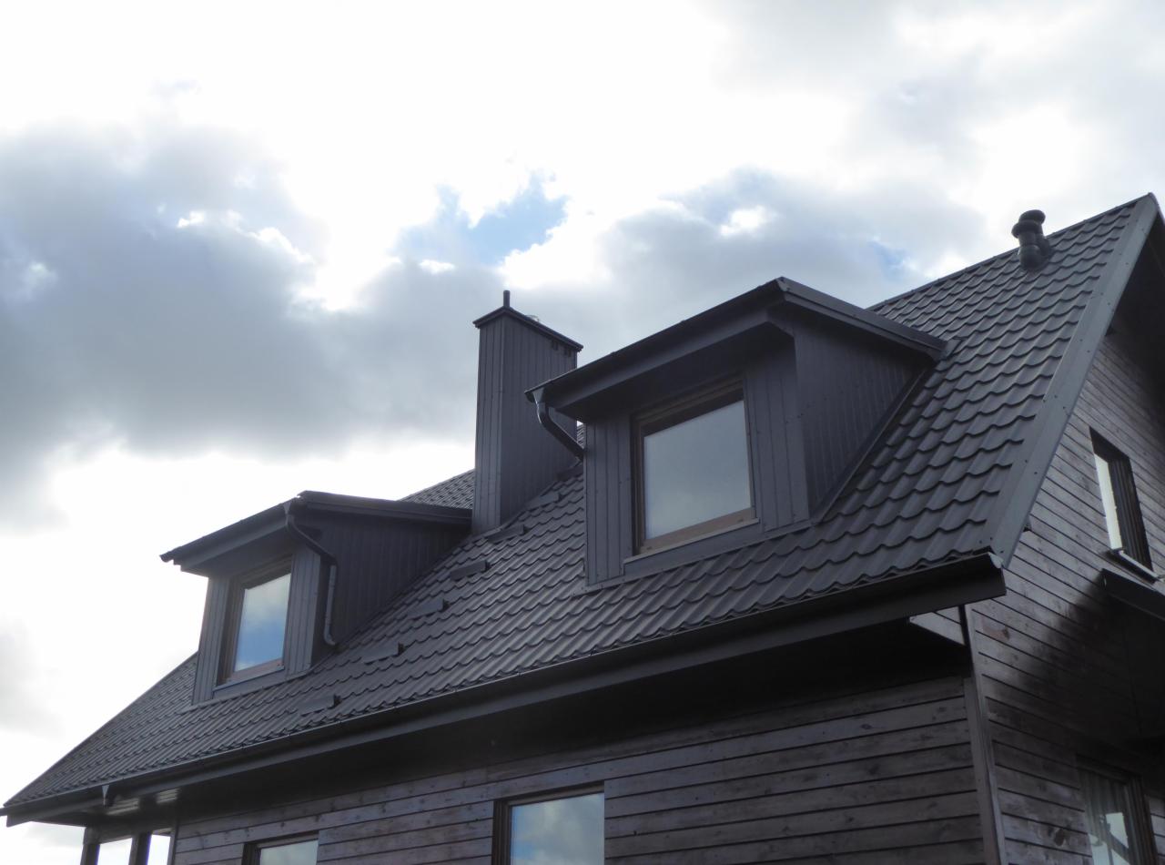 Dom z ciemnym dachem pokrytym dachówką bitumiczną, widoczne okna dachowe i komin na tle pochmurnego nieba.